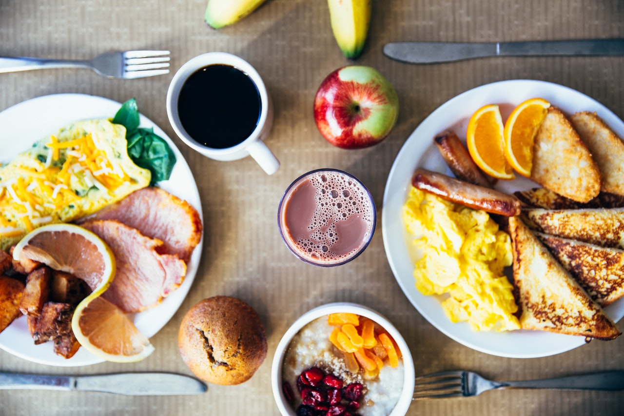 Plate of ham, hashbrowns, squash with cheese on left side of table. Plate of sausages, French toast, and scrambles eggs on right side. Oatmeal on bottom middle of table.