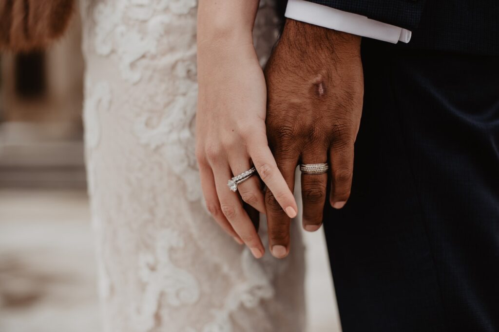 Hands of bride and groom slightly touching, both wearing silver colored wedding bands.