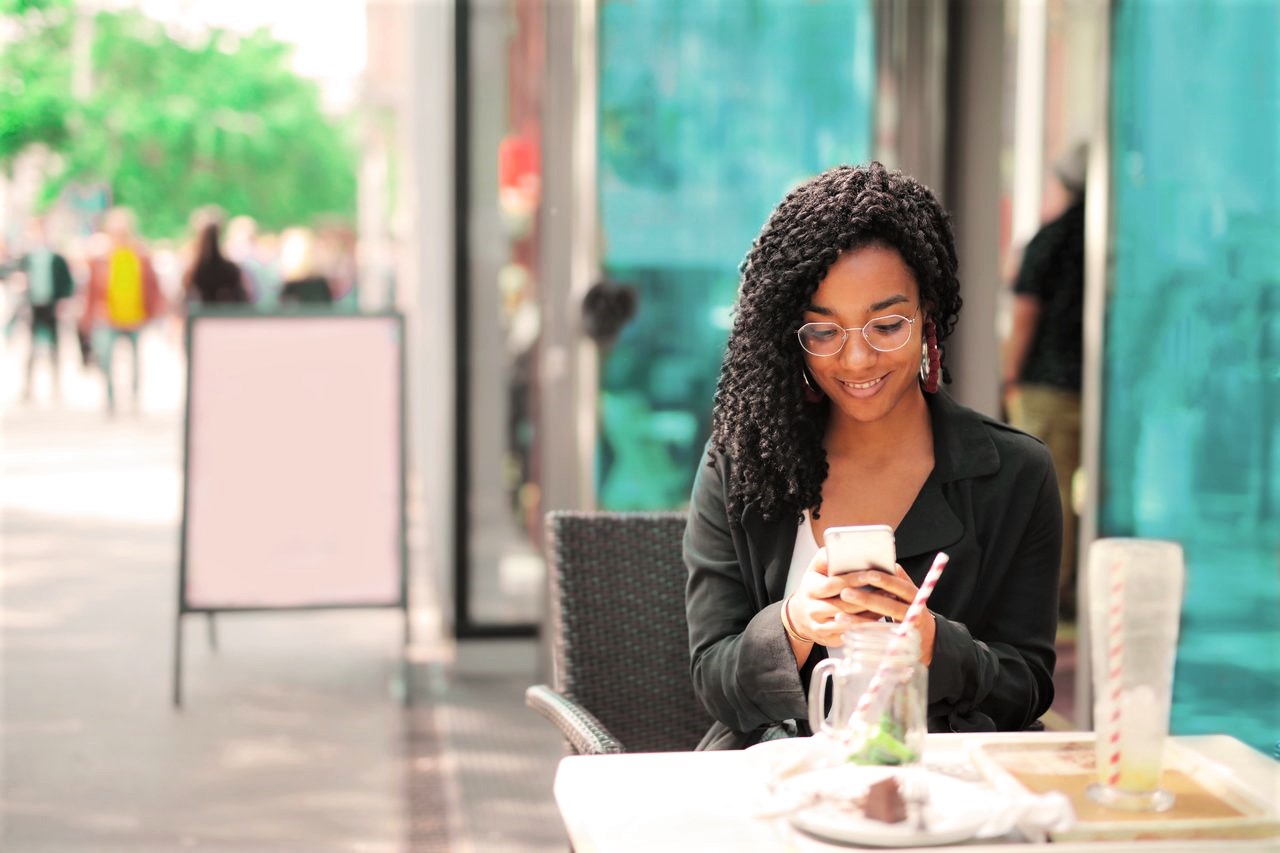 Black woman sitting outside on restaurant patio, wearing black long sleeve shirt holding her cellphone while not social distancing.