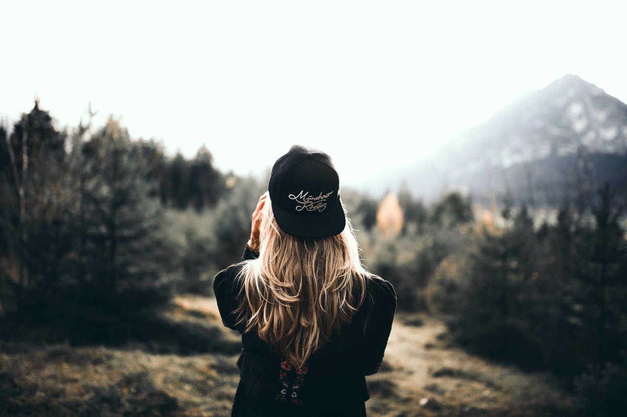 Back of blonde haired woman wearing black ball cap backwards and black sweatshirt, facing trees and mountain.