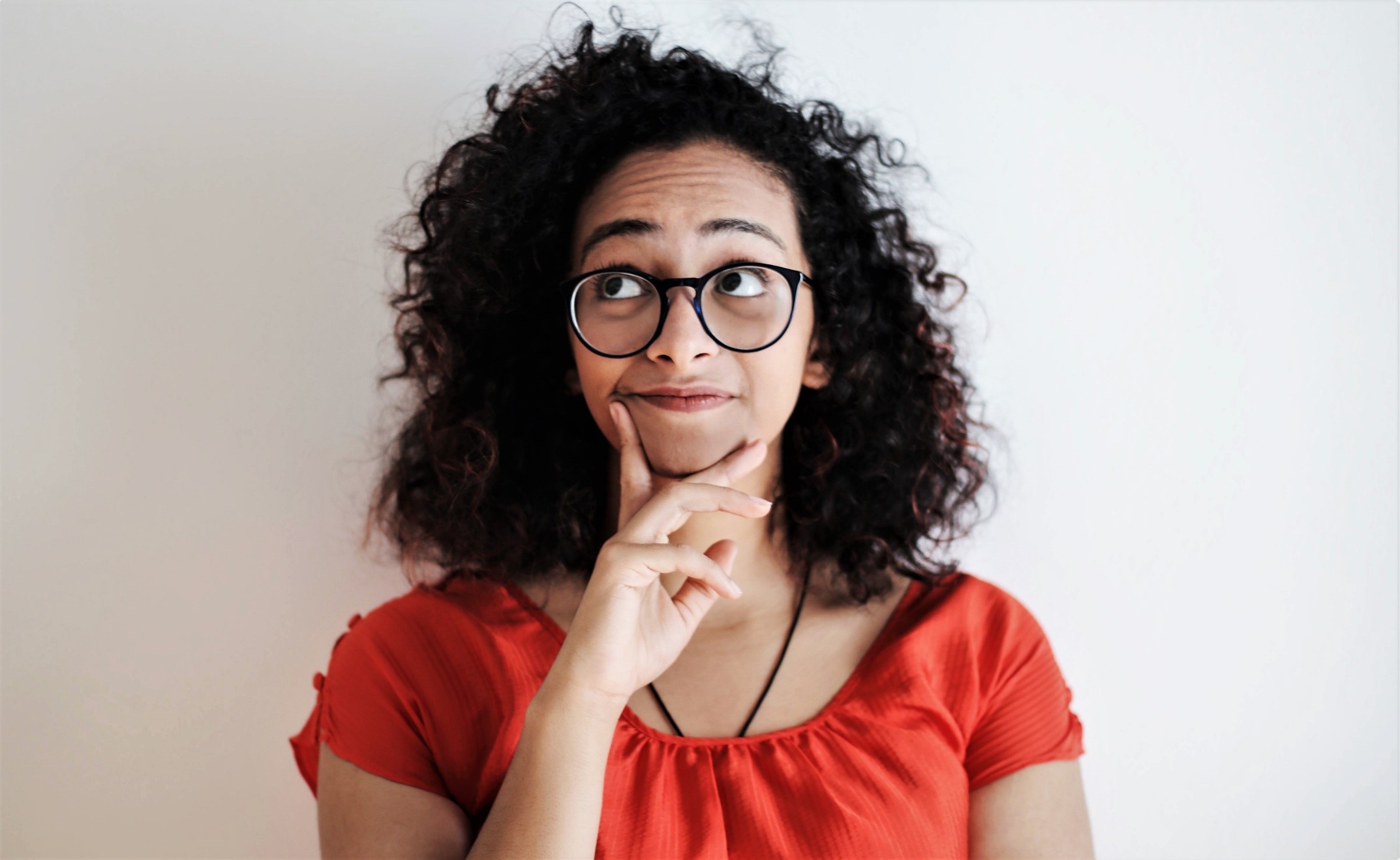 Young ethnic woman wearing red t-shirt, holding chin with one hand, thinking about how to say no