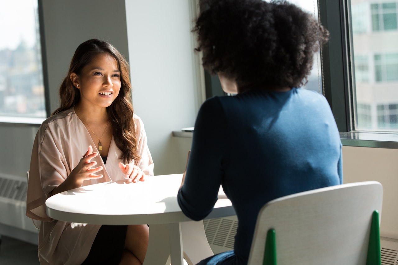 Asian woman wearing white top sitting across from Black woman figuring out how to say no. 