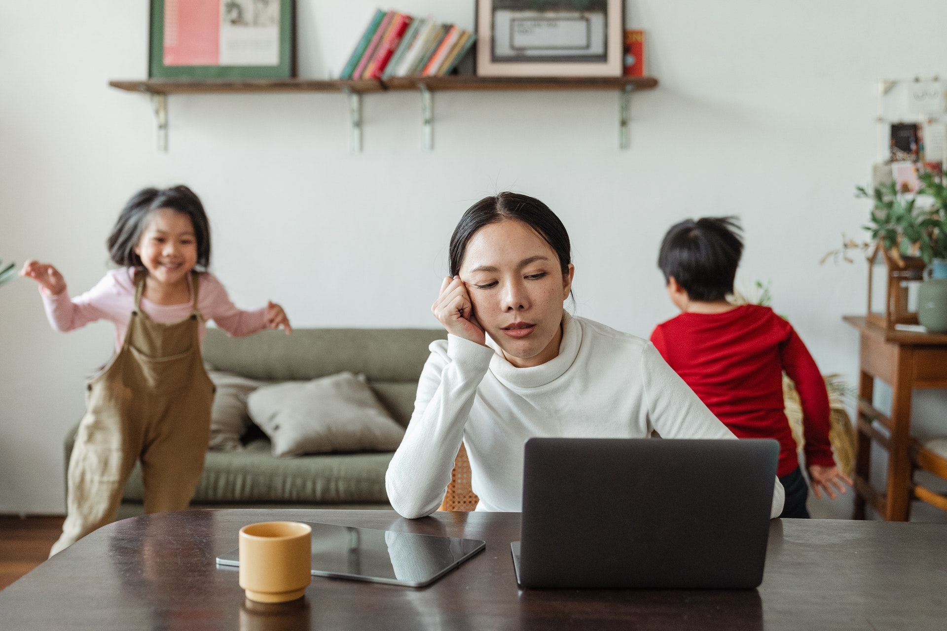 Asian mother sitting at laptop in living room experiencing burnout as daughter and son run in background.