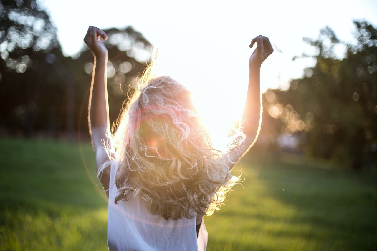 Back of a blonde woman with both her hands raised while standing in a field, full of gratitude.