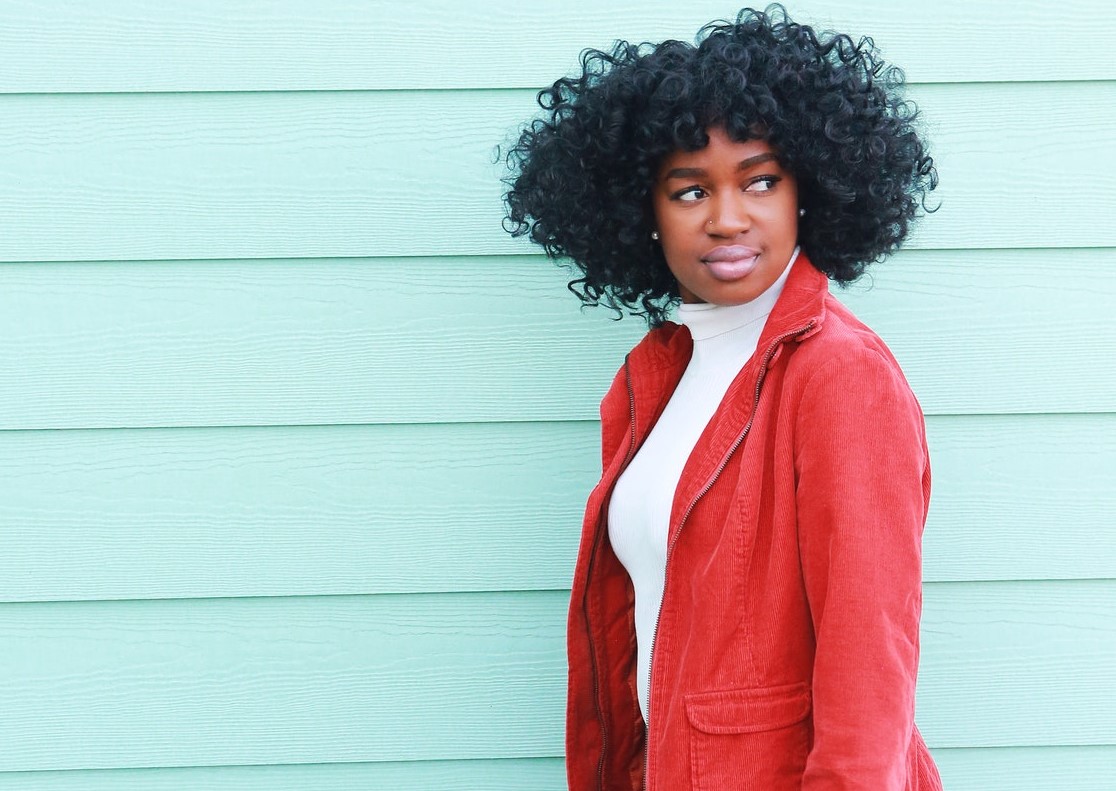 Black woman wearing red jacket, white turtleneck, and black pants reflecting on her identity.