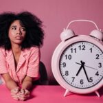 Young Black woman staring at large pink alarm clock procrastinating.