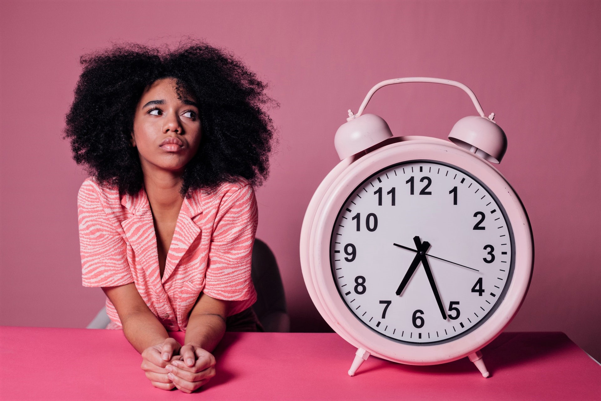 Young Black woman staring at large pink alarm clock procrastinating.