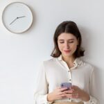 Young white business woman staring at her phone attempting time management while standing against white wall with analog clock hanging on wall.