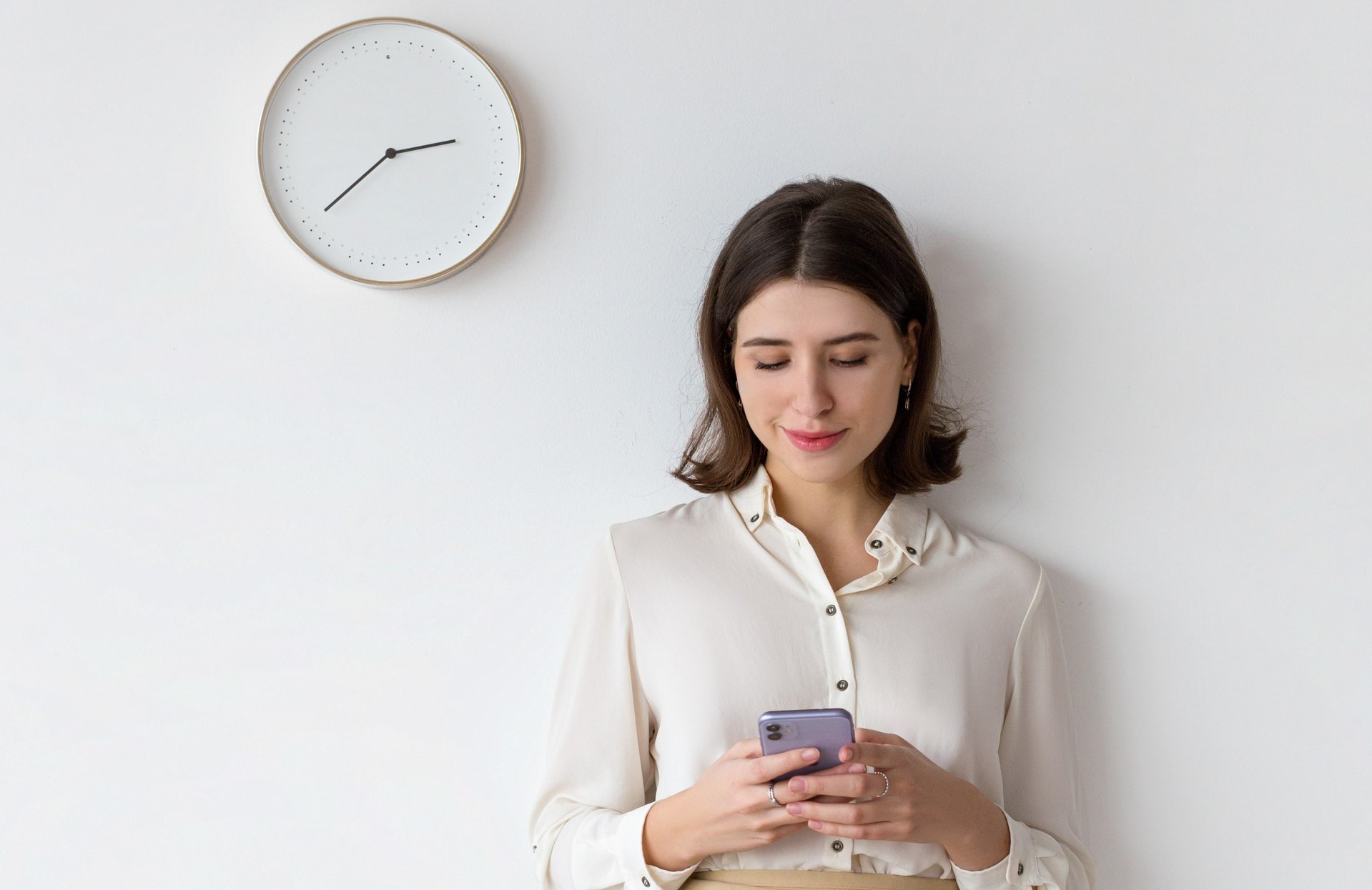 Young white business woman staring at her phone attempting time management while standing against white wall with analog clock hanging on wall.